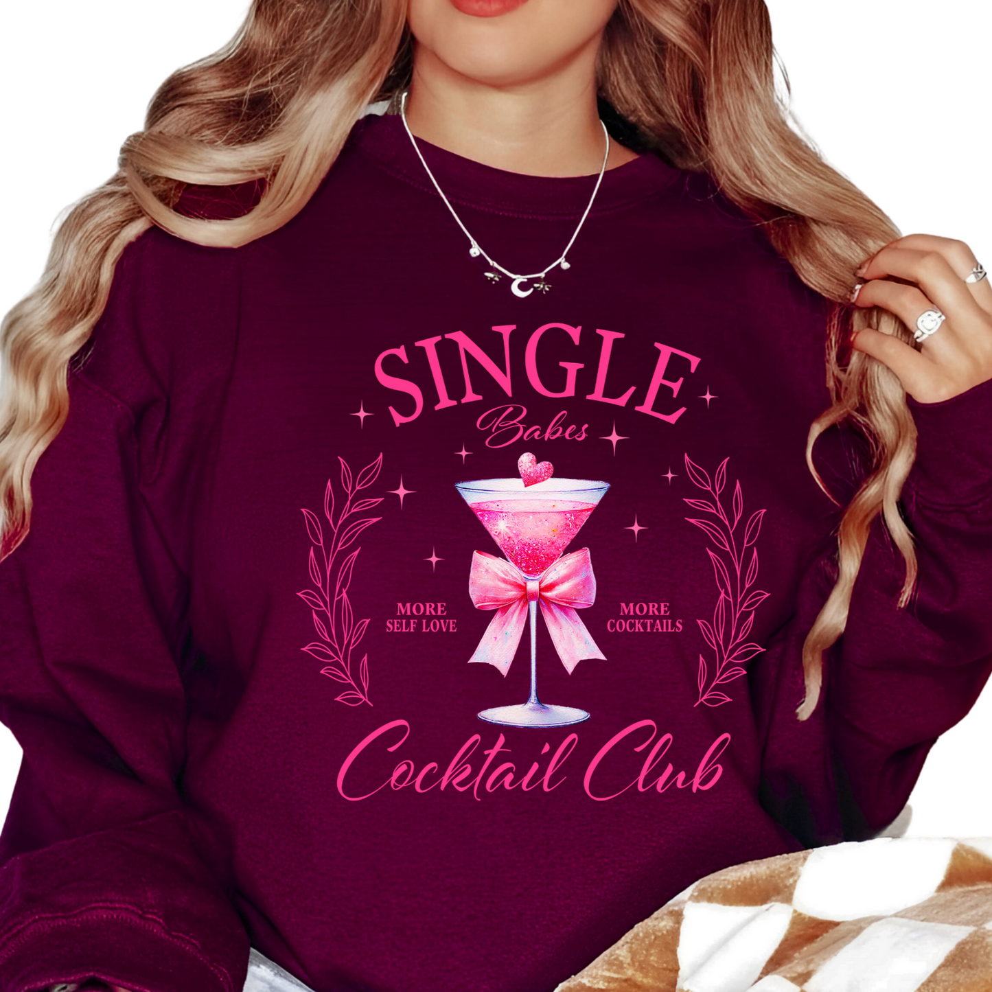 A woman sitting while wearing a black long-sleeve sweatshirt with text and a pink bow on it, indicating a playful and romantic theme appropriate for Valentine's Day.
