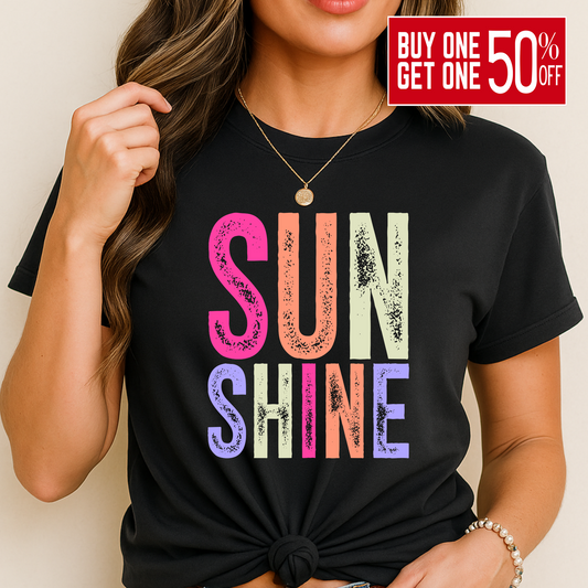 Woman wearing a bright “SUNSHINE” graphic T-shirt in Black, smiling in a relaxed summer setting.