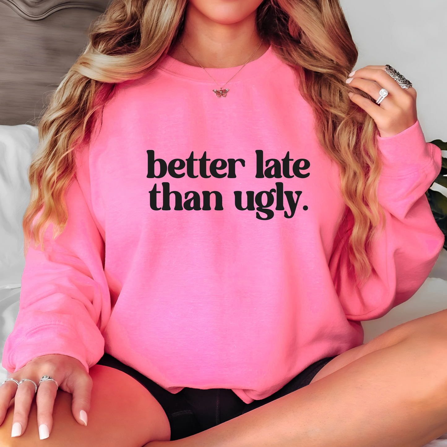 Bright pink fleece sweatshirt featuring “Better Late Than Ugly” in bold black lettering. Modeled by woman with long curled hair, accessorized with rings and necklace. Unisex fit for teens and women who love cozy fashion and sassy humor.