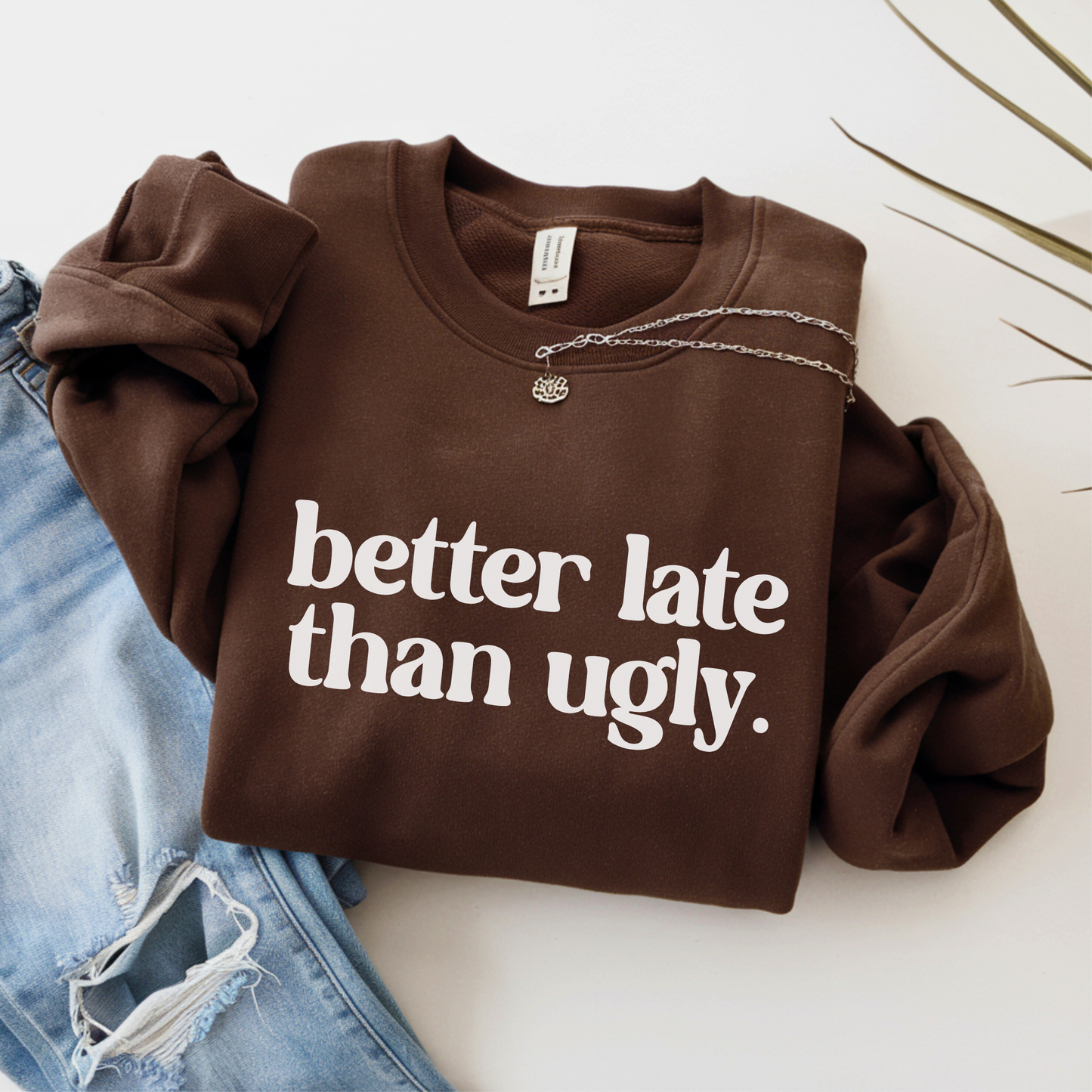 Brown sweatshirt with "Better Late Than Ugly" in white text, laid flat with rolled sleeves and silver necklace. Styled for women and teens, paired with distressed jeans. Cozy and cute unisex pullover for bold everyday wear.