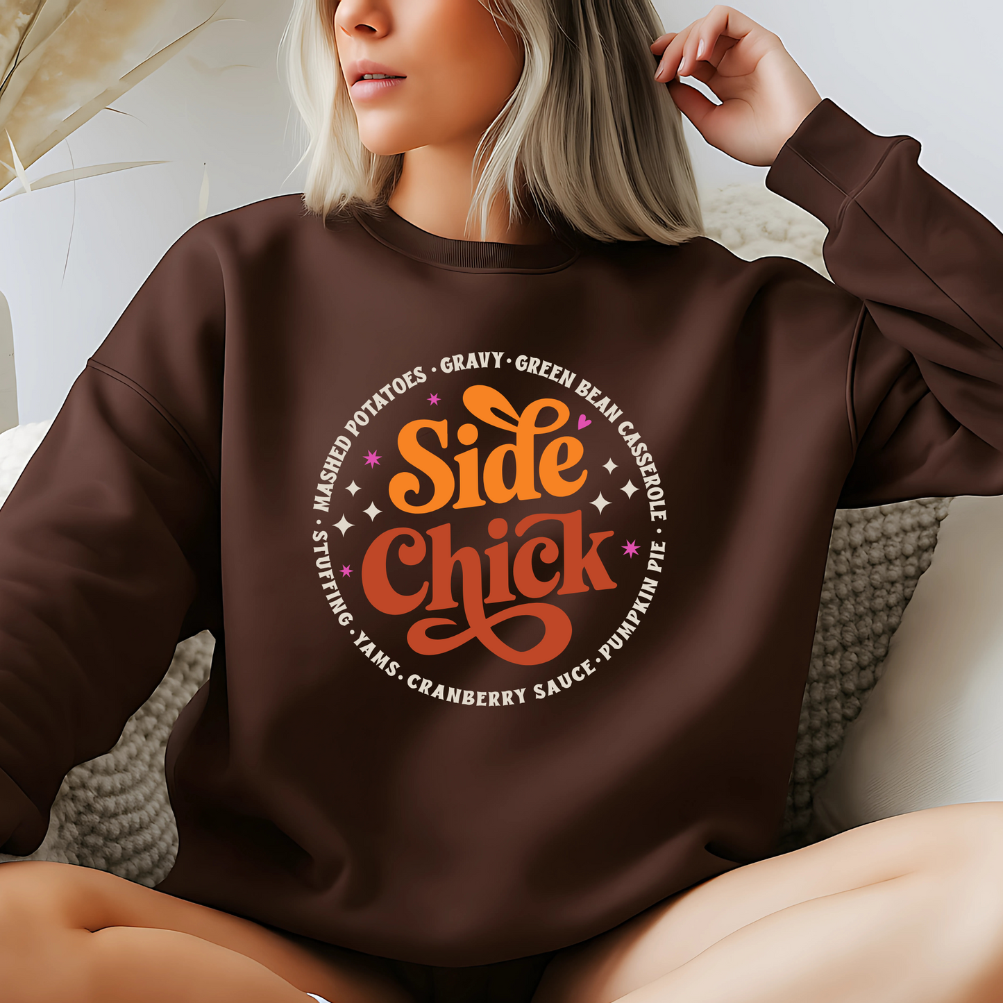 Dark brown “Side Chick” sweatshirt worn by woman lounging indoors. Cozy Thanksgiving sweater with retro lettering, perfect for fall loungewear, autumn celebrations, and funny holiday apparel. Great for foodies and side dish lovers.