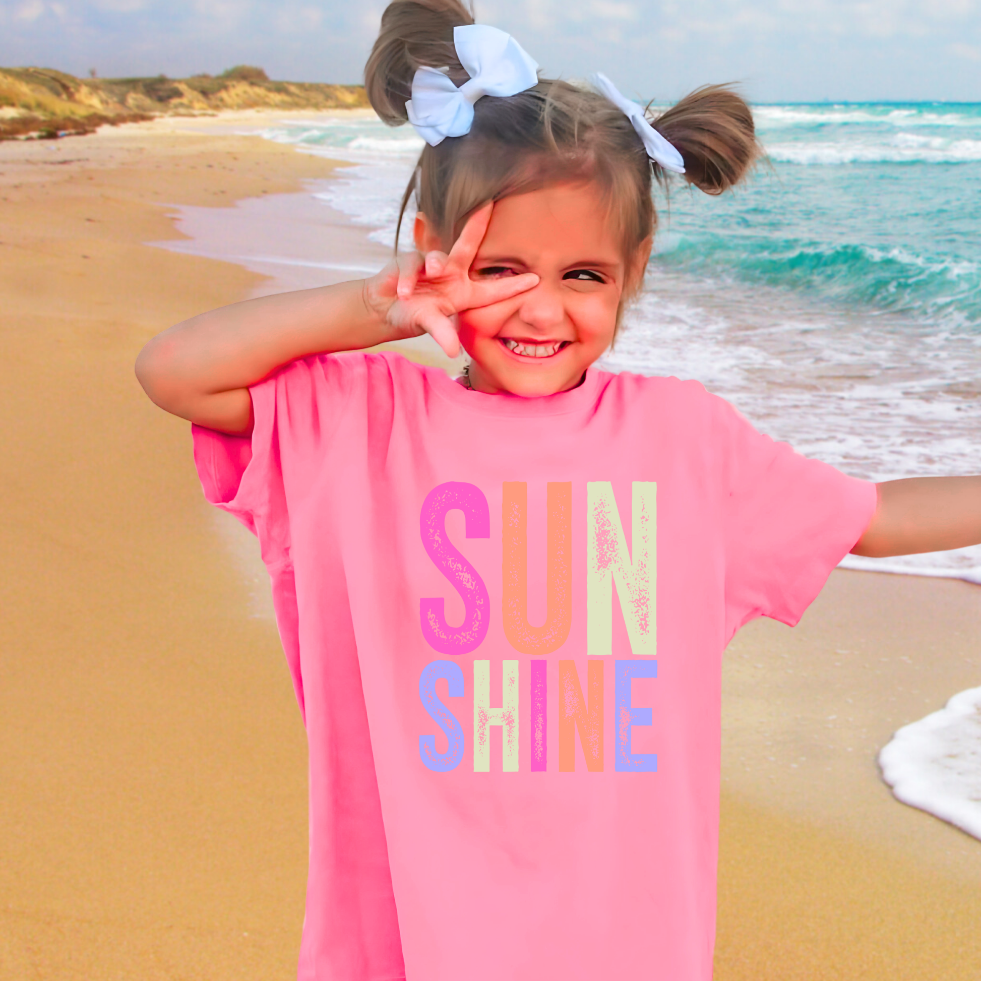 Girl on beach wearing bright pink “Sunshine” tee, smiling and posing