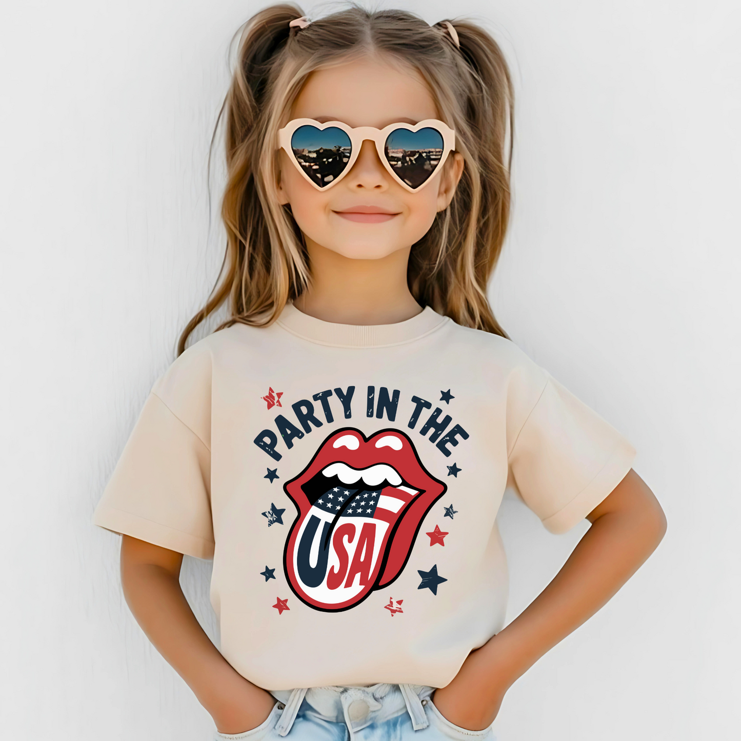 Stylish young girl wearing beige “Party in the USA” T-shirt with patriotic lips and American flag tongue design. Sporting heart-shaped sunglasses and denim shorts, posed against a white background. Perfect for 4th of July, Memorial Day, and summer celebrations with USA pride.