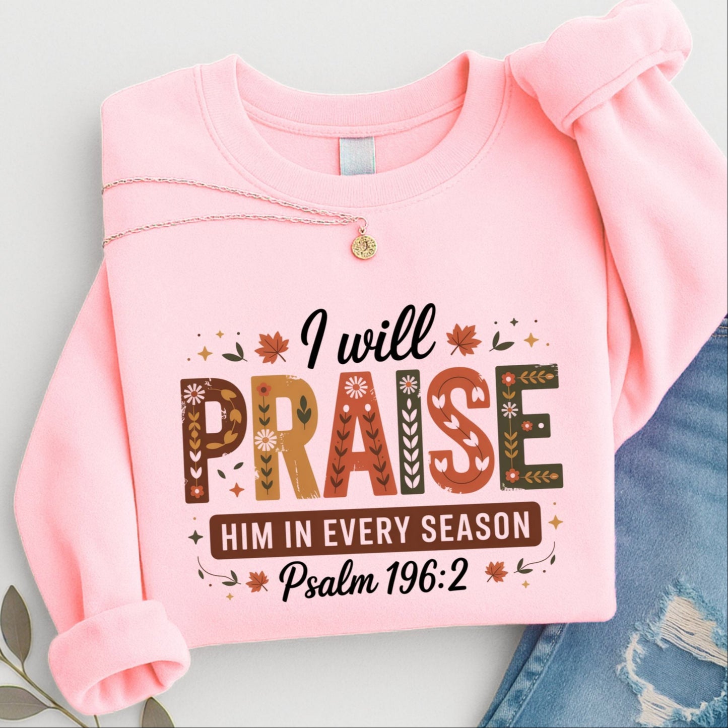 Folded pink “I Will Praise Him In Every Season” sweatshirt with fall floral lettering and Bible verse quote