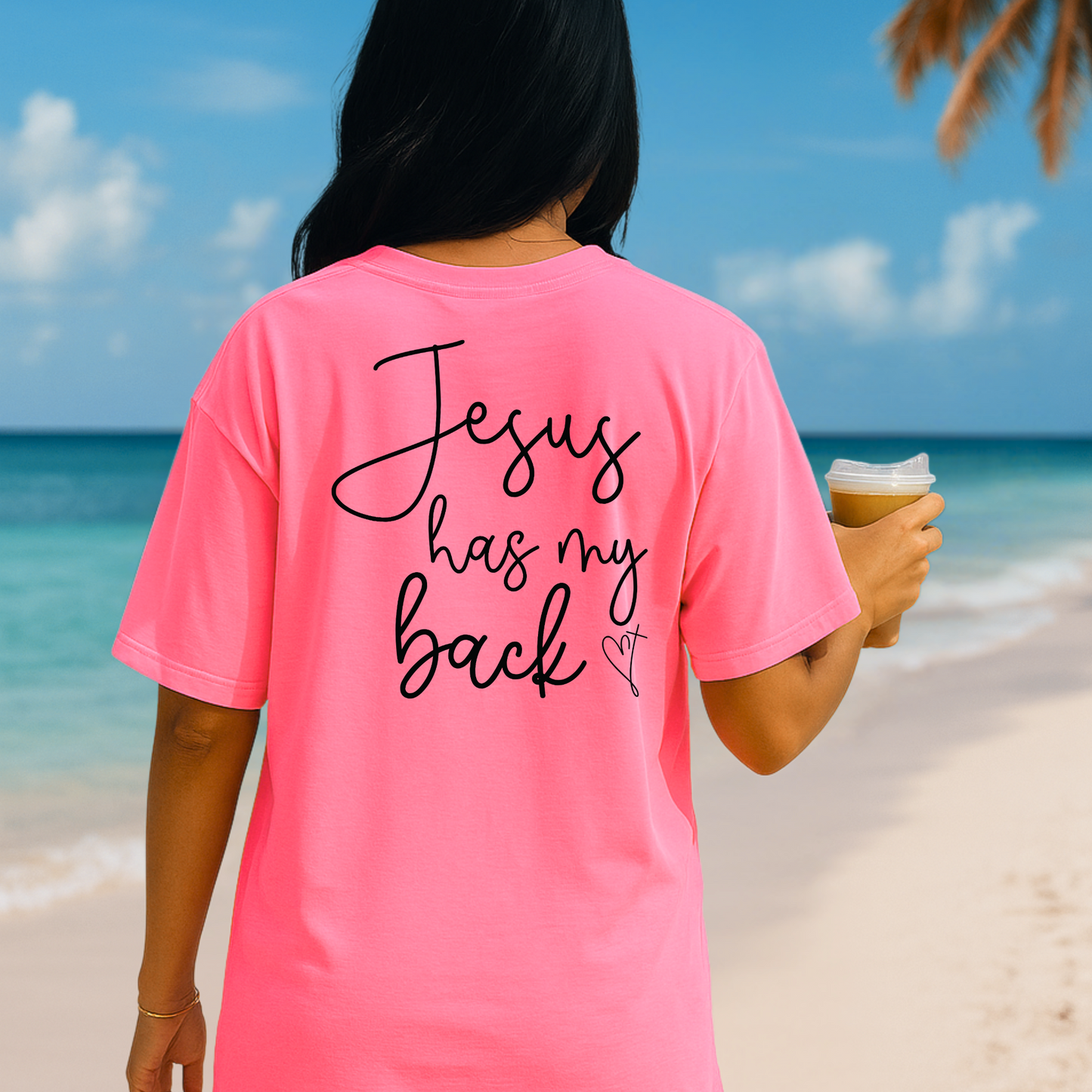 Woman wearing a pink “Jesus Has My Back” Christian T-shirt, back view, standing on a tropical beach holding iced coffee – inspirational women’s faith shirt.