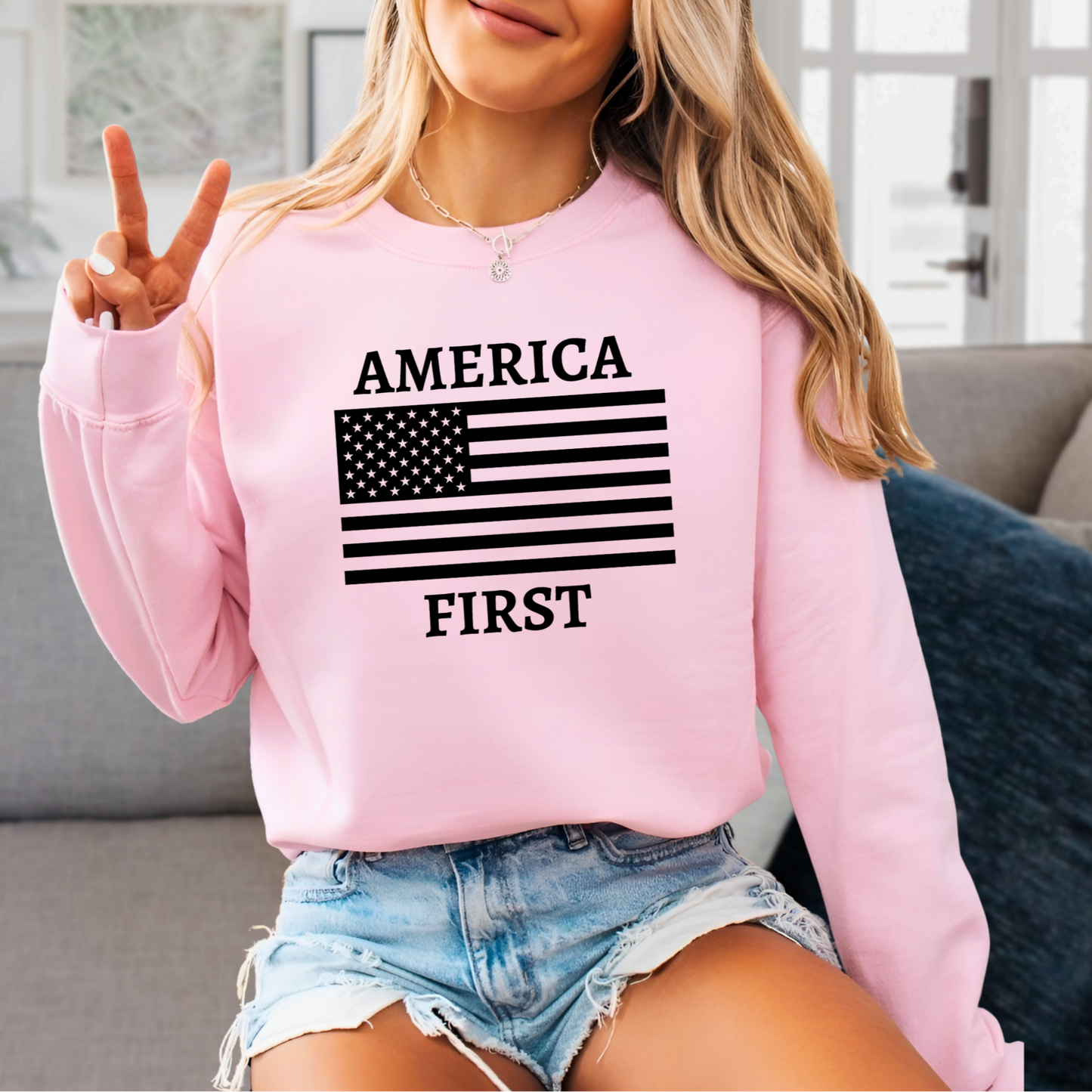 Woman giving the "Peace" sign while wearing a light Pink "America First" sweatshirt with a American flag.