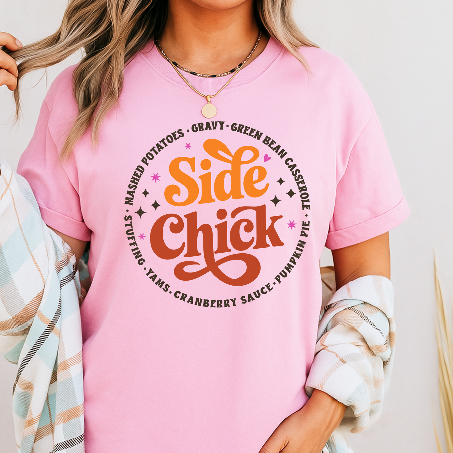 Model in a light pink “Side Chick” t-shirt styled with plaid cardigan. Fun and festive graphic print celebrating Thanksgiving side dishes in retro lettering. Great for teen girls, women, or moms wanting a playful and bright fall look. Adorable for Friendsgiving and casual November events.
