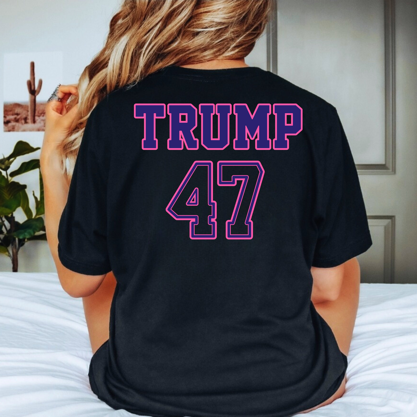 Woman wearing black Trump 47 shirt with Make America Trump Again front print, sitting on bed