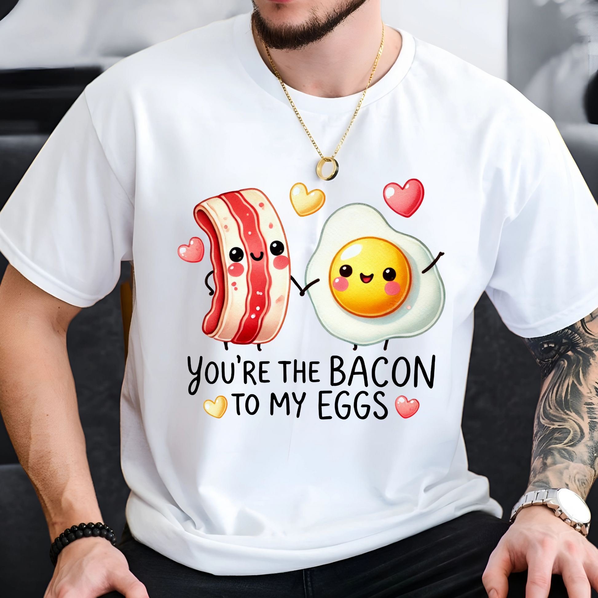 Man wearing a white T-shirt with cute cartoon bacon and egg holding hands and surrounded by hearts. The shirt reads 'You're the bacon to my eggs.'"