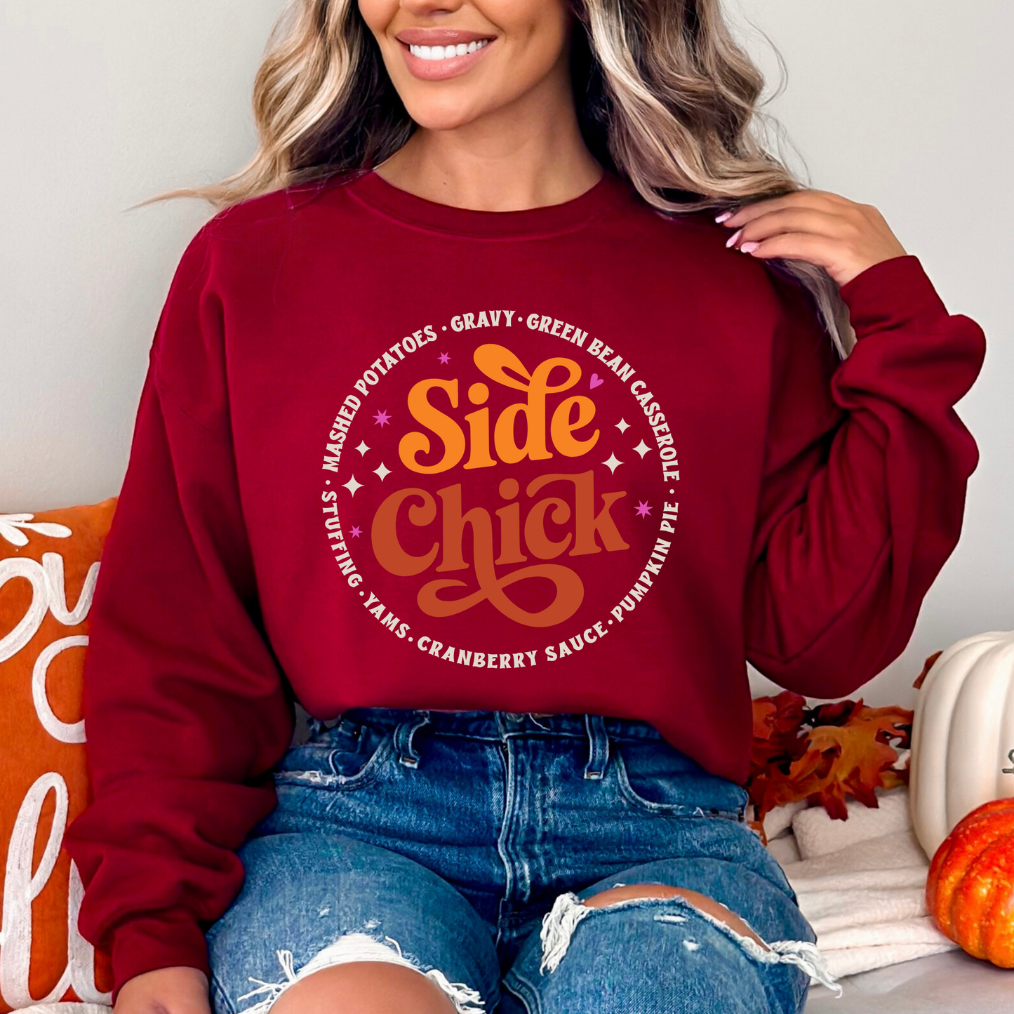 Woman in maroon “Side Chick” Thanksgiving sweatshirt paired with ripped denim shorts and pumpkins. Cozy fall pullover for women with a retro design celebrating side dishes. Perfect for autumn, holiday dinners, and cute seasonal outfits.