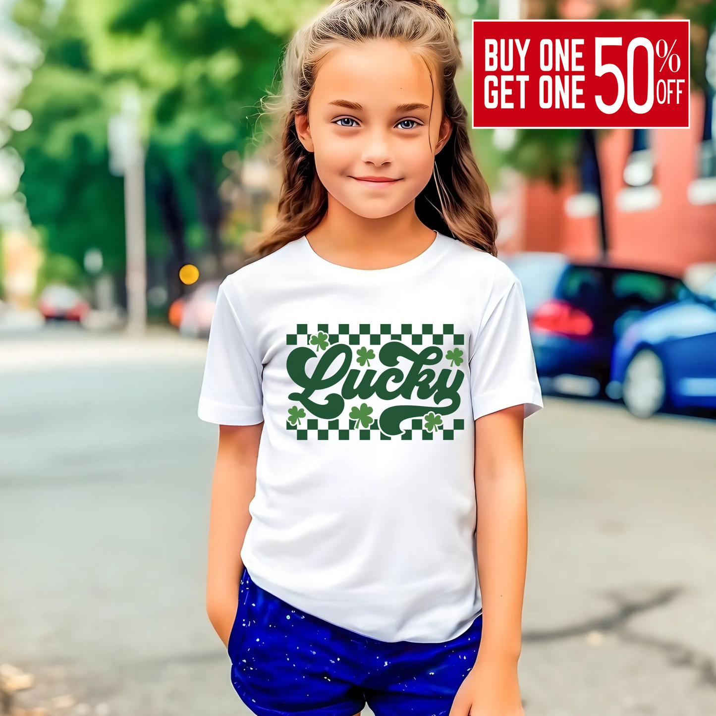 White T-shirt with 'Lucky' in green cursive, surrounded by shamrocks and a checkered pattern – perfect for St. Patrick’s Day.