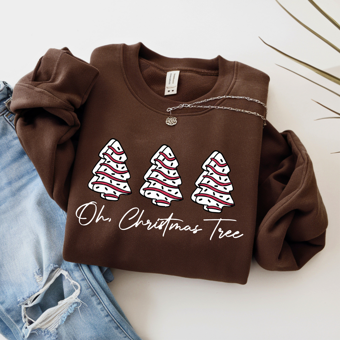 Flat lay of a brown Christmas sweatshirt featuring three festive red and white trees with “Oh Christmas Tree” text. Styled with ripped denim and silver necklace.