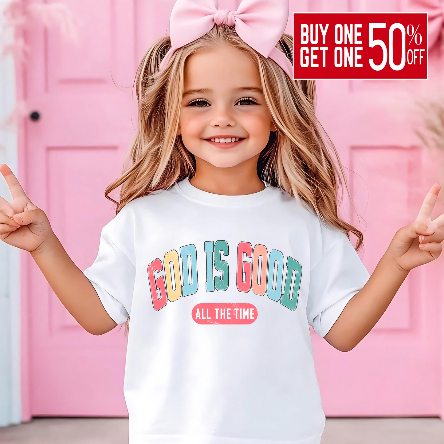 Happy young girl wearing a white 'God Is Good – All the Time' Easter t-shirt, posing with peace signs in front of a pink Easter-themed background