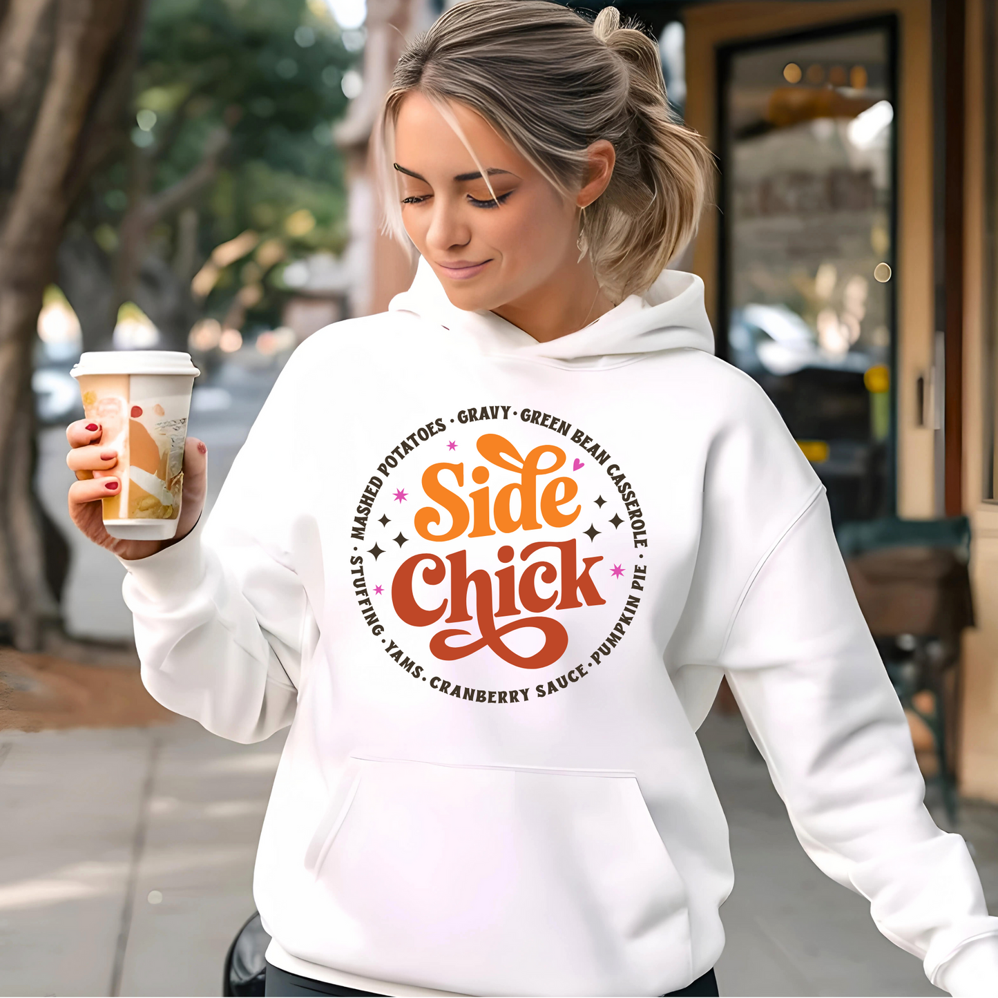 Woman holding coffee and wearing white “Side Chick” hoodie outside a cafe. Cute and cozy Thanksgiving hoodie with a food-themed design—perfect for holiday errands or pumpkin spice vibes.