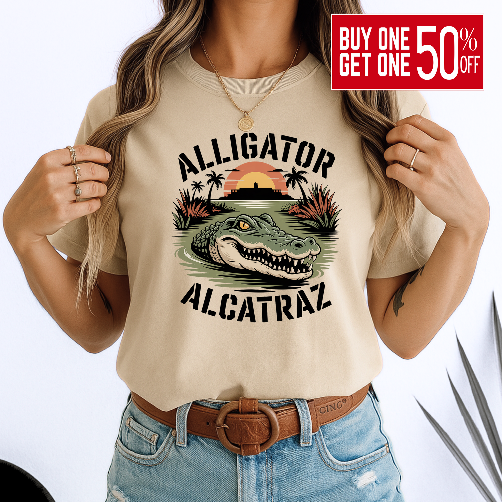 Woman in a beige Alligator Alcatraz T-shirt with vivid gator and sunset design. Soft unisex cotton tee styled with denim, great for travel or casual days.