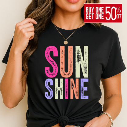 Woman wearing a bright “SUNSHINE” graphic T-shirt in Black, smiling in a relaxed summer setting.