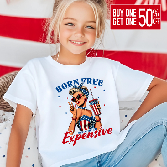 Child wearing a white t-shirt with 'Born Free But Now It's Expensive' graphic design in front of an American flag.