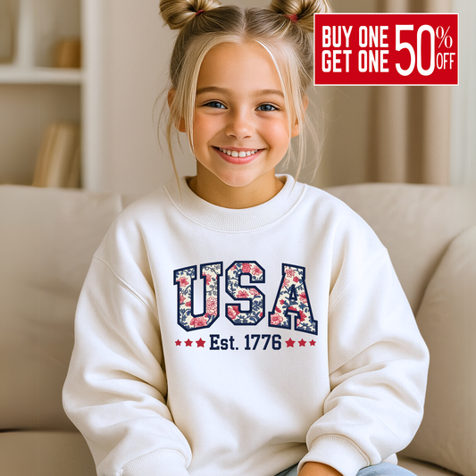 Girl in white USA Est. 1776 crewneck, seated and smiling on a beige couch.

