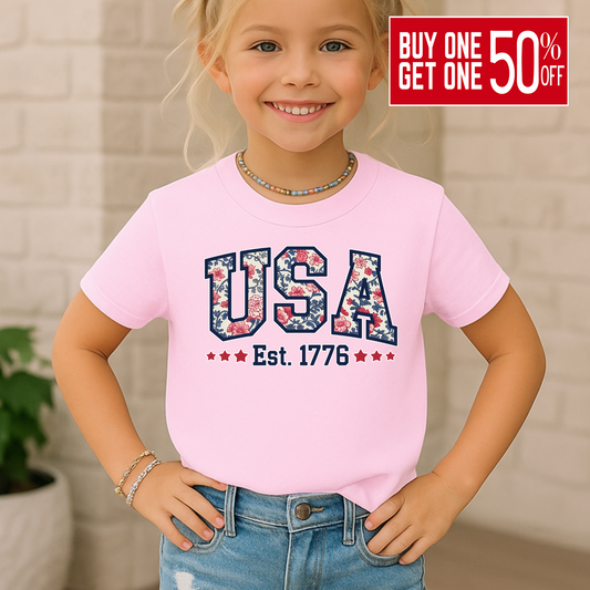 Girl wearing a light pink USA Est. 1776 youth t-shirt, smiling confidently in a casual outdoor  setting.

