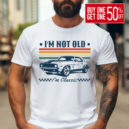 A bearded man wearing a white "I'm Not Old – I'm Classic" T-shirt with a retro muscle car design and vintage racing stripes in orange, yellow, and green. The graphic tee has a bold, nostalgic feel and is perfect for men who love cars and classic style. Great gift idea for car enthusiasts and birthdays.