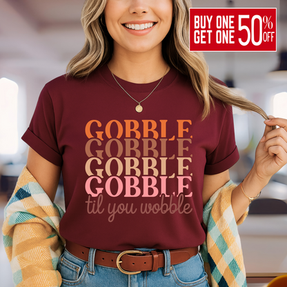 Smiling woman in a maroon shirt with fall design text “Gobble Til You Wobble,” layered with a cozy plaid cardigan – great for Friendsgiving