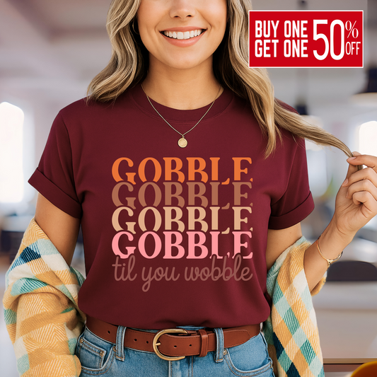 Smiling woman in a maroon shirt with fall design text “Gobble Til You Wobble,” layered with a cozy plaid cardigan – great for Friendsgiving