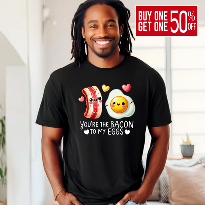 Smiling man wearing a black T-shirt with kawaii-style bacon and egg holding hands, surrounded by colorful hearts. Design says 'You're the bacon to my eggs