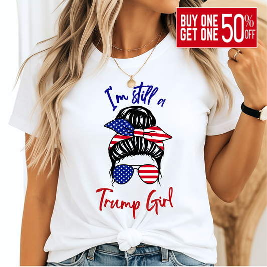 Woman wearing white “I’m Still a Trump Girl” patriotic T-shirt with USA flag bun and sunglasses design.
