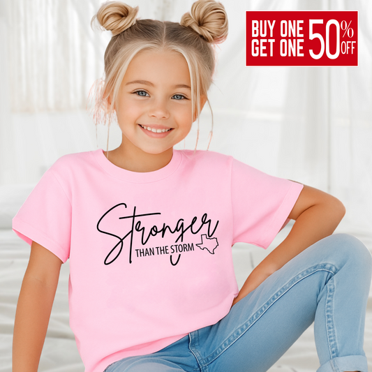 Young girl wearing light pink “Stronger Than the Storm” Texas kids T-shirt, smiling in bright room.

